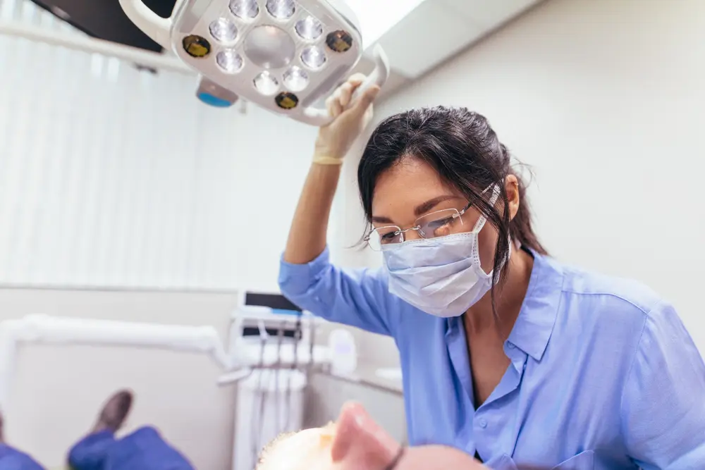 Female dentist wearing mask adjusting light with patient on chair at dental clinic. Female doctor preparing for a dental treatment on male patient.