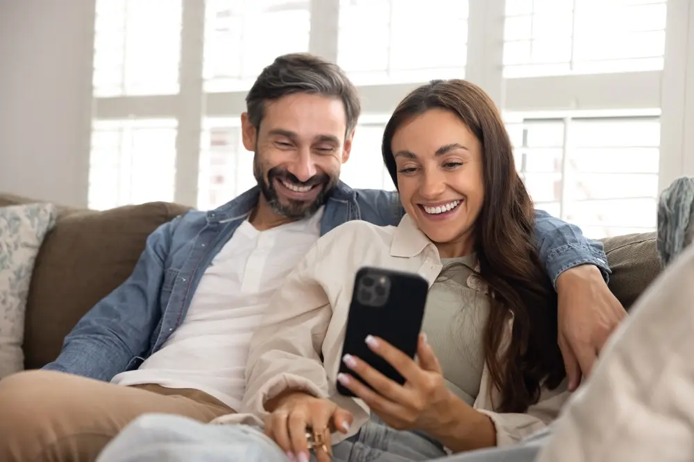Smiling young couple looking at a smartphone laughing together on the couch