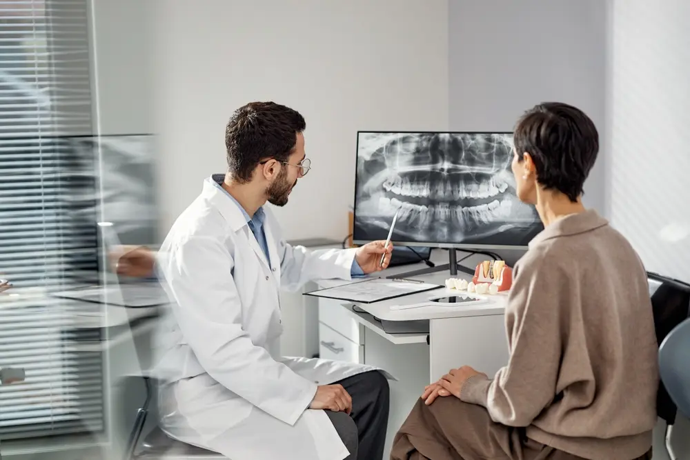 Young male dentist explaining dental X-ray results to middle-aged Caucasian woman patient in dental office, dentist using pen to indicate details on monitor, patient listening attentively