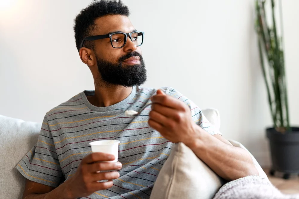 Man with beard and glasses eating yogurt on sofa, relaxing and contemplating in a bright home environment